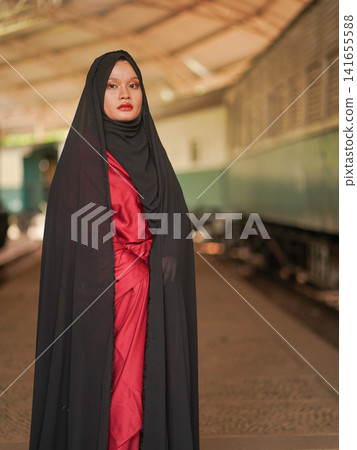 Portrait of a beautiful young Muslim woman wearing an elegant black abaya and red dress posing confidently in a rustic industrial warehouse, modern modest fashion concept. Portrait of a beautiful young Muslim woman wearing an elegant black abaya and red dress posing confidently in a rustic industrial warehouse, modern modest fashion concept. 141655588