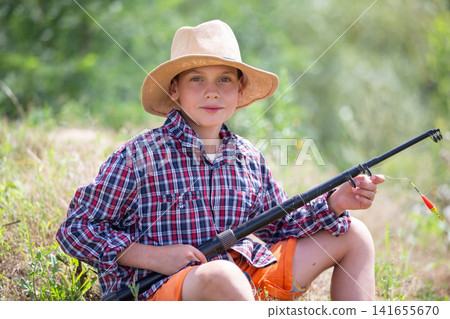 A cheerful boy dressed in a checkered shirt and a straw hat sits with a fishing rod, smiling at the camera. The setting features a vibrant green riverbank and bright sunlight. A cheerful boy dressed in a checkered shirt and a straw hat sits with a fishing rod, smiling at the camera. The setting features a vibrant green riverbank and bright sunlight. 141655670