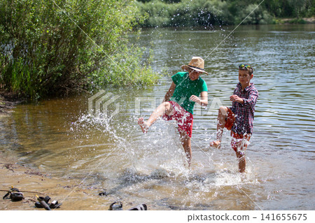 Two children splash water playfully at the river's edge during a sunny summer afternoon in a natural setting surrounded by greenery Two children splash water playfully at the river's edge during a sunny summer afternoon in a natural setting surrounded by greenery 141655675