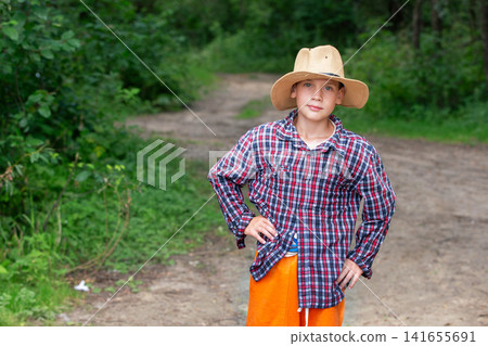 Young boy in a straw hat stands proudly on a forest path surrounded by greenery during daytime Young boy in a straw hat stands proudly on a forest path surrounded by greenery during daytime 141655691