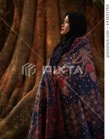 Portrait of a beautiful young Muslim woman wearing an elegant black hijab and traditional red Jambi Batik, posing confidently in a rustic industrial warehouse, modern modest fashion concept. Portrait of a beautiful young Muslim woman wearing an elegant black hijab and traditional red Jambi Batik, posing confidently in a rustic industrial warehouse, modern modest fashion concept. 141655768