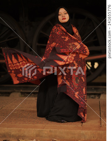Portrait of a beautiful young Muslim woman wearing an elegant black hijab and traditional red Jambi Batik, posing confidently in a rustic industrial warehouse, modern modest fashion concept. 141655776