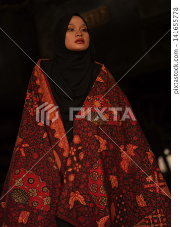 Portrait of a beautiful young Muslim woman wearing an elegant black hijab and traditional red Jambi Batik, posing confidently in a rustic industrial warehouse, modern modest fashion concept. Portrait of a beautiful young Muslim woman wearing an elegant black hijab and traditional red Jambi Batik, posing confidently in a rustic industrial warehouse, modern modest fashion concept. 141655778
