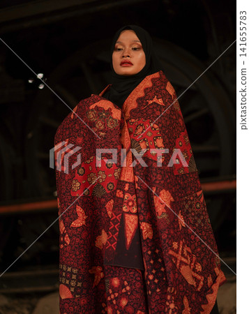 Portrait of a beautiful young Muslim woman wearing an elegant black hijab and traditional red Jambi Batik, posing confidently in a rustic industrial warehouse, modern modest fashion concept. Portrait of a beautiful young Muslim woman wearing an elegant black hijab and traditional red Jambi Batik, posing confidently in a rustic industrial warehouse, modern modest fashion concept. 141655783