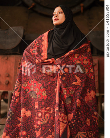 Beautiful Indonesian Muslim woman wearing a black hijab and traditional red Jambi Batik dress, posing confidently in an industrial warehouse setting. Beautiful Indonesian Muslim woman wearing a black hijab and traditional red Jambi Batik dress, posing confidently in an industrial warehouse setting. 141655904