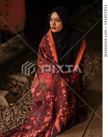 Beautiful Indonesian Muslim woman wearing a black hijab and traditional red Jambi Batik dress, posing confidently in an industrial warehouse setting. Beautiful Indonesian Muslim woman wearing a black hijab and traditional red Jambi Batik dress, posing confidently in an industrial warehouse setting. 141655913