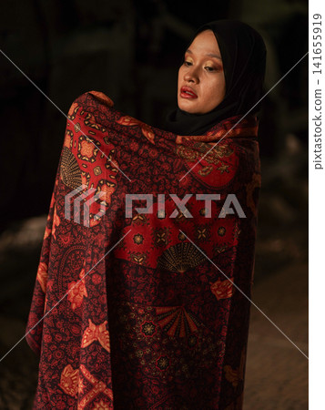 Beautiful Indonesian Muslim woman wearing a black hijab and traditional red Jambi Batik dress, posing confidently in an industrial warehouse setting. Beautiful Indonesian Muslim woman wearing a black hijab and traditional red Jambi Batik dress, posing confidently in an industrial warehouse setting. 141655919