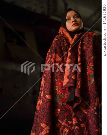 Beautiful Indonesian Muslim woman wearing a black hijab and traditional red Jambi Batik dress, posing confidently in an industrial warehouse setting. Beautiful Indonesian Muslim woman wearing a black hijab and traditional red Jambi Batik dress, posing confidently in an industrial warehouse setting. 141655920
