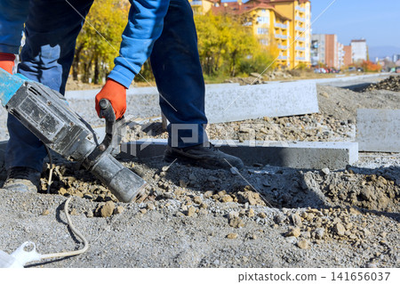 Worker in gloves operates jackhammer on construction site while preparing renovation road. Worker in gloves operates jackhammer on construction site while preparing renovation road. 141656037