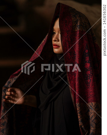 Confident young Asian woman showcasing modest fashion in a red Jambi Batik dress and black hijab, posing in a rustic warehouse environment. Confident young Asian woman showcasing modest fashion in a red Jambi Batik dress and black hijab, posing in a rustic warehouse environment. 141656302