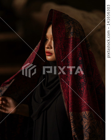 Confident young Asian woman showcasing modest fashion in a red Jambi Batik dress and black hijab, posing in a rustic warehouse environment. Confident young Asian woman showcasing modest fashion in a red Jambi Batik dress and black hijab, posing in a rustic warehouse environment. 141656303