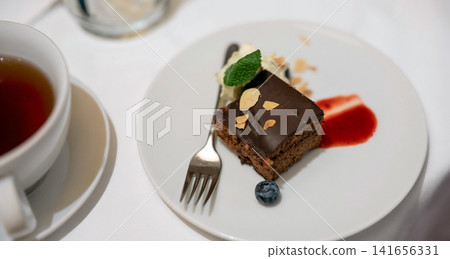 A delicious slice of chocolate cake topped with almonds, cream, mint, and a blueberry, served on a white plate next to a cup of tea, perfect for a gourmet dessert. A delicious slice of chocolate cake topped with almonds, cream, mint, and a blueberry, served on a white plate next to a cup of tea, perfect for a gourmet dessert. 141656331