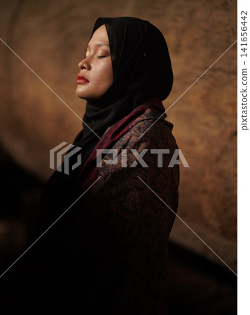 Confident young Asian woman showcasing modest fashion in a red Jambi Batik dress and black hijab, posing in a rustic warehouse environment. Confident young Asian woman showcasing modest fashion in a red Jambi Batik dress and black hijab, posing in a rustic warehouse environment. 141656442