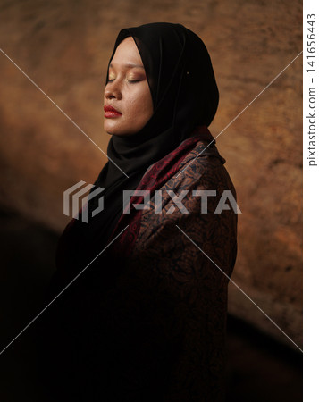 Confident young Asian woman showcasing modest fashion in a red Jambi Batik dress and black hijab, posing in a rustic warehouse environment. 141656443