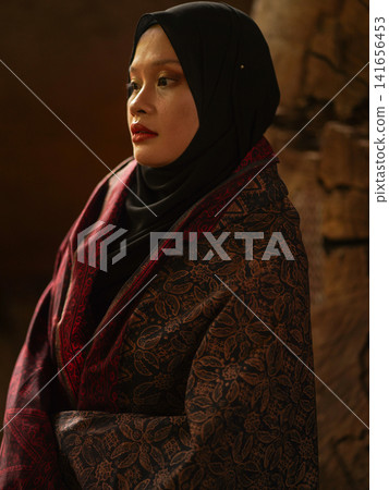 Confident young Asian woman showcasing modest fashion in a red Jambi Batik dress and black hijab, posing in a rustic warehouse environment. Confident young Asian woman showcasing modest fashion in a red Jambi Batik dress and black hijab, posing in a rustic warehouse environment. 141656453