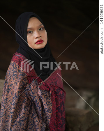 A fashionable young woman wearing a traditional black hijab and an elegant long red dress standing confidently against a weathered, vintage green door. Perfect for modest fashion, cultural, and lifest A fashionable young woman wearing a traditional black hijab and an elegant long red dress standing confidently against a weathered, vintage green door. Perfect for modest fashion, cultural, and lifest 141656621