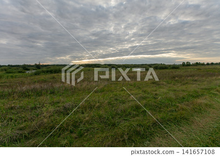 Cloudy sky above a tranquil field near water at sunset in a rural landscape Cloudy sky above a tranquil field near water at sunset in a rural landscape 141657108