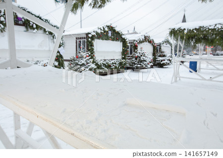 Snow covers a market with decorated stalls and trees in a town square during winter Snow covers a market with decorated stalls and trees in a town square during winter 141657149