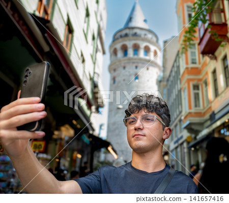 Istanbul, turkey, august 2, 2025. Young man holding smartphone for selfie while visiting galata tower, capturing travel memories in city street Istanbul, turkey, august 2, 2025. Young man holding smartphone for selfie while visiting galata tower, capturing travel memories in city street 141657416