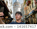 Istanbul, turkey, august 2, 2025. Young man using smartphone to capture a selfie, showing the iconic galata tower in the background while traveling in istanbul, turkey 141657417