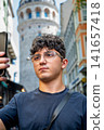 Istanbul, turkey, august 2, 2025. Young man wearing glasses holding a smartphone in front of galata tower, exploring istanbul as a tourist 141657418