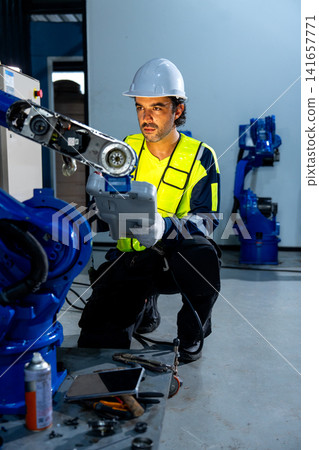 Engineer in yellow vest and white helmet adjusts blue robotic arm with focused intensity in clean industrial workshop Engineer in yellow vest and white helmet adjusts blue robotic arm with focused intensity in clean industrial workshop 141657771