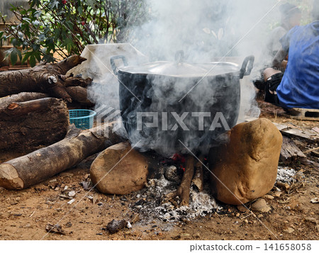 A scene of traditional Thai rural cooking featuring a steaming metal pot on a primitive firewood stove made of stones. Showing smoke, ashes, and a local person in the background, representing authenti 141658058