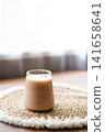 Homemade iced coffee prepared with frozen cherry juice ice cubes in a clear glass. Refreshing summer drink served on a natural woven placemat with soft daylight and minimal home interior background. 141658641