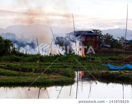 Controlled field burning near a village on Lake Inle, Myanmar, with flames, smoke, and water reflections framed by mountains at dusk Controlled field burning near a village on Lake Inle, Myanmar, with flames, smoke, and water reflections framed by mountains at dusk 141658820