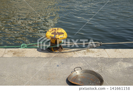 Weathered yellow mooring bollard with nautical rope and empty paella pan on harbor pier 141661158