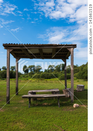 Rustic wooden picnic shelter in grassy meadow with scattered clouds Rustic wooden picnic shelter in grassy meadow with scattered clouds 141661519