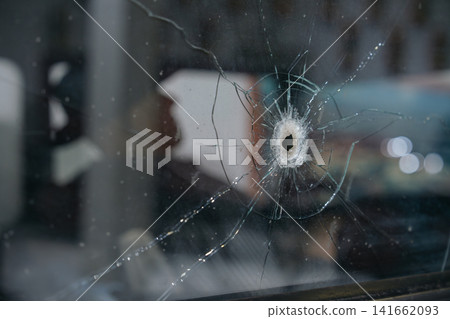 Detailed close up of a bullet hole surrounded by cracked glass on a historic vehicle window. The image highlights impact damage and shattered glass textures. Detailed close up of a bullet hole surrounded by cracked glass on a historic vehicle window. The image highlights impact damage and shattered glass textures. 141662093