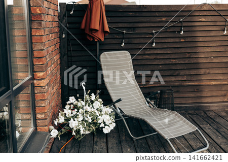 Modern outdoor terrace or patio with lounge chair, blooming white flowers and wooden privacy fence. Cozy backyard relaxation space at a contemporary home, showcasing comfort and stylish living. Modern outdoor terrace or patio with lounge chair, blooming white flowers and wooden privacy fence. Cozy backyard relaxation space at a contemporary home, showcasing comfort and stylish living. 141662421