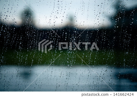Raindrops on window glass during rainfall with blurred natural landscape background. Rainy weather, climate change, storm, precipitation and seasonal weather concept. Natural environmental pattern. Raindrops on window glass during rainfall with blurred natural landscape background. Rainy weather, climate change, storm, precipitation and seasonal weather concept. Natural environmental pattern. 141662424
