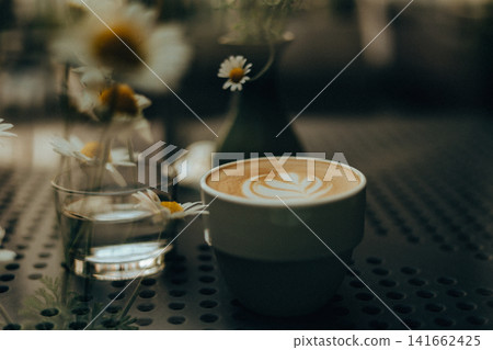 A beautifully crafted latte with heart-shaped latte art sits on a perforated metal cafe table beside a glass of water and delicate chamomile flowers. A beautifully crafted latte with heart-shaped latte art sits on a perforated metal cafe table beside a glass of water and delicate chamomile flowers. 141662425