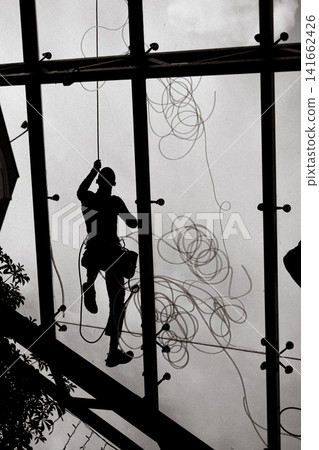 Silhouette of an industrial rope access worker cleaning or maintaining a glass facade. High-contrast urban scene of safety, labor, architecture. 141662426