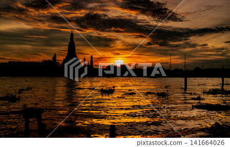 Silhouette of Wat Arun at dramatic sunset over the Chao Phraya River in Bangkok, Thailand, glowing reflections, clouds and iconic Buddhist temple architecture. Famous historic Buddhist landmark. 141664026