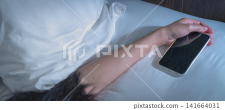 Asian woman sleeping in bed at home and hand holding mobile phone. Woman using smartphone in bedroom. Young woman addicted using smartphone. Texting on cellphone with tired until falling asleep. Asian woman sleeping in bed at home and hand holding mobile phone. Woman using smartphone in bedroom. Young woman addicted using smartphone. Texting on cellphone with tired until falling asleep. 141664031