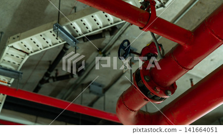 Red fire protection sprinkler piping network suspended from a modern building ceiling, featuring industrial engineering, fire suppression equipment, safety infrastructure and mechanical installation. Red fire protection sprinkler piping network suspended from a modern building ceiling, featuring industrial engineering, fire suppression equipment, safety infrastructure and mechanical installation. 141664051