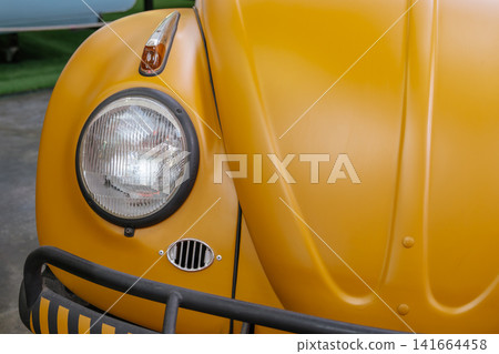 A close-up view of the headlight and curved hood of a yellow vintage Beetle automobile. The image highlights classic automotive styling, smooth body lines, and retro design details. A close-up view of the headlight and curved hood of a yellow vintage Beetle automobile. The image highlights classic automotive styling, smooth body lines, and retro design details. 141664458