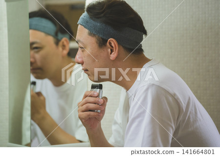 一位四十多歲的男子正在用電動刮鬍刀刮鬍子,側面照在鏡子裡,映出他的身影。 一位四十多歲的男子正在用電動刮鬍刀刮鬍子,側面照在鏡子裡,映出他的身影。 141664801