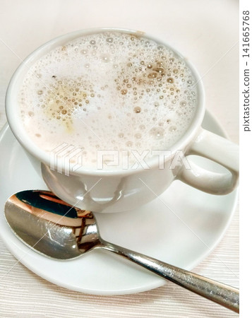 Close-up of cappuccino in white cup with foam and spoon on saucer, elegant table setting Close-up of cappuccino in white cup with foam and spoon on saucer, elegant table setting 141665768