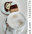 Coffee cup with frothy milk beside plate of chocolate and vanilla cake slices on white table 141665770