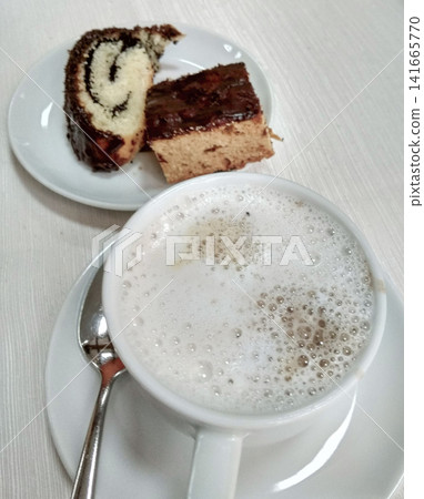 Coffee cup with frothy milk beside plate of chocolate and vanilla cake slices on white table Coffee cup with frothy milk beside plate of chocolate and vanilla cake slices on white table 141665770