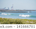 Waves crashing onto the sandy beach and gantry cranes in the harbor visible in the distance. 141666701