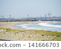 A view of a sandy beach lush with coastal plants and a harbor with large cranes lined up in the distance. 141666703