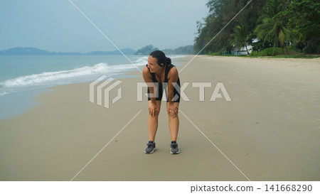 Young woman in black sportswear resting after sprinting along a sandy beach with lush green tropical vegetation and hills clearly visible behind. Active outdoor concept Young woman in black sportswear resting after sprinting along a sandy beach with lush green tropical vegetation and hills clearly visible behind. Active outdoor concept 141668290