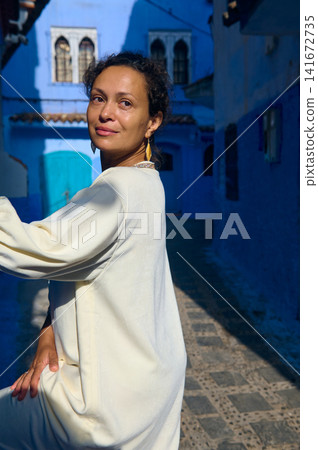 Woman in white dress posing in blue city alley of Chefchaouen, Morocco 141672735