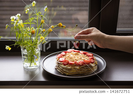 Homemade layered crepe cake topped with fresh strawberries on a plate by a window. Cozy summer breakfast scene with wildflowers, natural light and a calm slow living atmosphere. Summertime dessert. Homemade layered crepe cake topped with fresh strawberries on a plate by a window. Cozy summer breakfast scene with wildflowers, natural light and a calm slow living atmosphere. Summertime dessert. 141672751