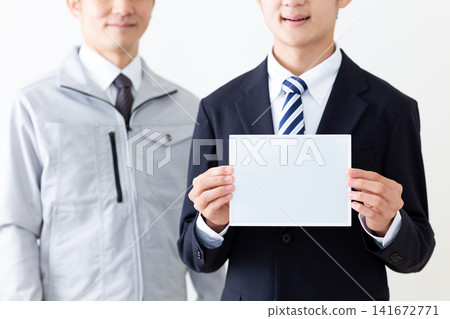 一位身穿西裝的年輕男子,手持一塊普通的白色白板;一位身穿工作服的中年男子。 一位身穿西裝的年輕男子,手持一塊普通的白色白板;一位身穿工作服的中年男子。 141672771